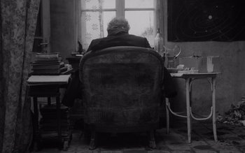 Movie still from “Satantango” (1994), directed by Béla Tarr – An older man sitting in a chair in front of a window; Medium shot, High angle