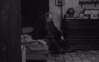 Movie still from “Satantango” (1994), directed by Béla Tarr – An older man sitting on a bed in a room; Wide shot, High angle