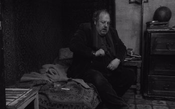Movie still from “Satantango” (1994), directed by Béla Tarr – An older man sitting on a bed with a cigarette in his hand; Medium shot, High angle