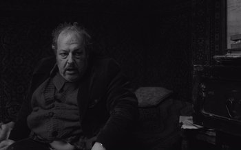 Movie still from “Satantango” (1994), directed by Béla Tarr – An older man sitting on a couch in a dark room; Medium shot, Low angle