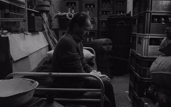 Movie still from “Satantango” (1994), directed by Béla Tarr – A man sitting on a bed in a cluttered room; Medium shot, High angle