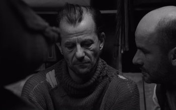 Movie still from “Satantango” (1994), directed by Béla Tarr – An older man wearing a sweater with a beard; Close Up shot, Over the shoulder angle