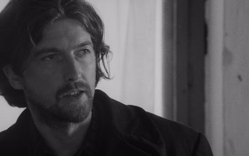 Movie still from “Satantango” (1994), directed by Béla Tarr – Black and white photograph of a man with long hair; Close Up shot, Over the shoulder angle