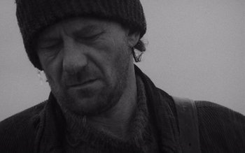 Movie still from “Satantango” (1994), directed by Béla Tarr – A black and white photo of a man in a hat and scarf; Close Up shot, Low angle