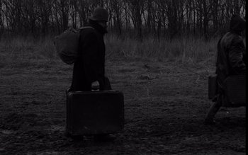 Movie still from “Satantango” (1994), directed by Béla Tarr – A man in a hat and a coat is holding a suitcase; Wide shot, High angle