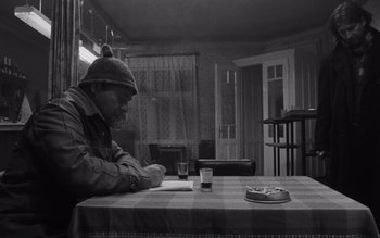 Movie still from “Satantango” (1994), directed by Béla Tarr – A man sitting at a table with a pizza on the table; Medium shot, High angle