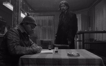 Movie still from “Satantango” (1994), directed by Béla Tarr – A man standing next to a table with a glass of beer; Medium shot, Over the shoulder angle