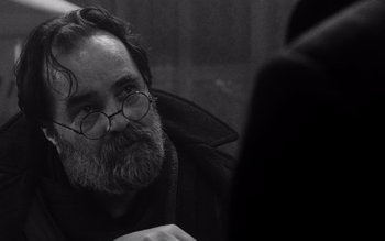 Movie still from “Satantango” (1994), directed by Béla Tarr – A man with a goatee wearing glasses and a black jacket; Close Up shot, Over the shoulder angle