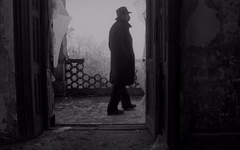 Movie still from “Satantango” (1994), directed by Béla Tarr – A man wearing a hat and a long coat walking through a door way; Wide shot, Low angle