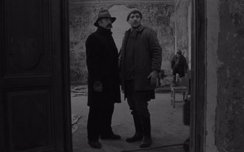 Movie still from “Satantango” (1994), directed by Béla Tarr – Two men standing next to each other in front of a wall; Medium shot, Low angle