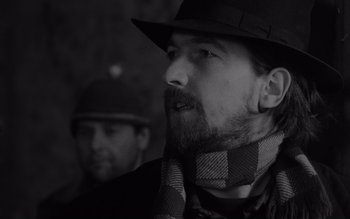 Movie still from “Satantango” (1994), directed by Béla Tarr – A man with a beard wearing a hat and a scarf; Close Up shot, Low angle