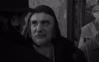 Movie still from “Satantango” (1994), directed by Béla Tarr – An old man with long hair and a beard wearing a hat; Close Up shot, Over the shoulder angle
