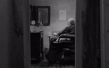 Movie still from “Satantango” (1994), directed by Béla Tarr – An older man sitting in a chair in a room; Wide shot, High angle