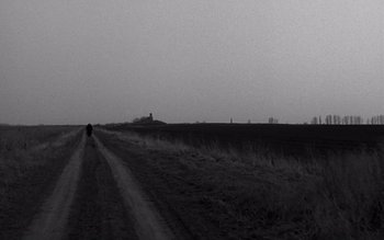 Movie still from “Satantango” (1994), directed by Béla Tarr – A person walking down a dirt road near a field; Extreme Wide shot, High angle