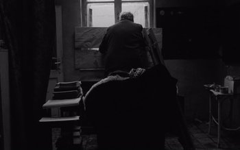 Movie still from “Satantango” (1994), directed by Béla Tarr – An older man sitting in front of a window; Wide shot, Low angle