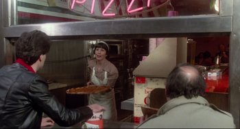 Movie still from “Saturday Night Fever” (1977), directed by John Badham – A woman holding a pan of pizza in front of a neon sign; Medium shot, Over the shoulder angle