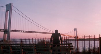 Movie still from “Saturday Night Fever” (1977), directed by John Badham – A man standing on top of a fence looking at a bridge; Extreme Wide shot, Low angle