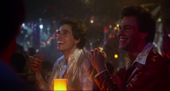 Movie still from “Saturday Night Fever” (1977), directed by John Badham – Two young men are laughing together at a party; Medium shot, Over the shoulder angle