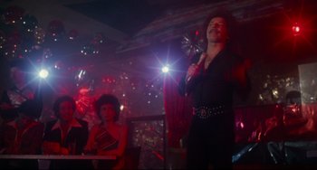 Movie still from “Saturday Night Fever” (1977), directed by John Badham – A man in a black shirt is holding a microphone; Wide shot, Low angle