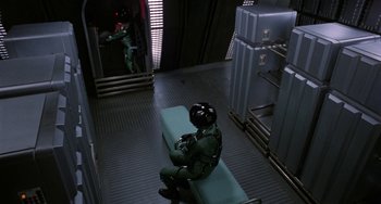 Movie still from “Saturn 3” (1980), directed by John Barry – A man in a green uniform sitting on a bench; Wide shot, Overhead angle