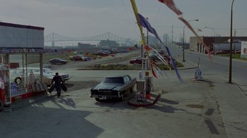 Movie still from “Scanners” (1981), directed by David Cronenberg – A car is parked in a parking lot next to gas pumps; Extreme Wide shot, High angle