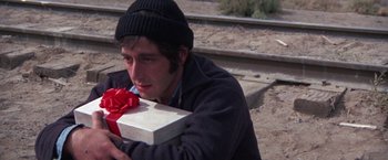 Movie still from “Scarecrow” (1973), directed by Jerry Schatzberg – A man holding a gift box on the side of the road; Close Up shot, High angle