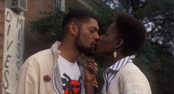 Movie still from “School Daze” (1988), directed by Spike Lee – A man and a woman kissing each other in front of a building; Medium shot, Low angle