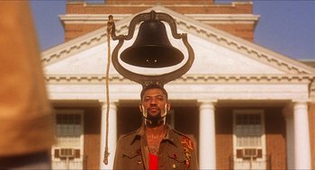 Movie still from “School Daze” (1988), directed by Spike Lee – A man standing in front of a building with a bell on top of his head; Medium shot, Low angle