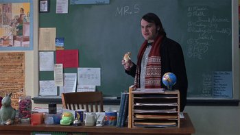 Movie still from “School of Rock” (2003), directed by Richard Linklater – A man standing in front of a chalkboard holding a sandwich; Medium shot, Over the shoulder angle