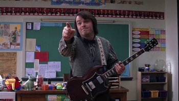 Movie still from “School of Rock” (2003), directed by Richard Linklater – A man holding a guitar in front of a chalkboard in a classroom; Medium shot, Low angle