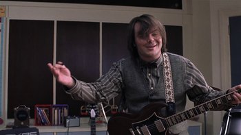Movie still from “School of Rock” (2003), directed by Richard Linklater – A man holding a guitar in a room with other guitars; Medium shot, Low angle