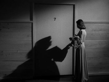 Movie still from “Secret Beyond the Door...” (1947), directed by Fritz Lang – A black and white photo of a woman holding a bouquet of flowers in front of a door; Wide shot, Low angle