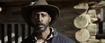 Movie still from “Godless” (2017), directed by Scott Frank – A man wearing a cowboy hat standing in front of a building; Close Up shot, Low angle