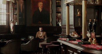 Movie still from “Secrets & Lies” (1996), directed by Mike Leigh – A man sitting in front of a large painting of a man; Wide shot, Over the shoulder angle