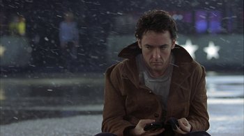 Movie still from “Serendipity” (2001), directed by Peter Chelsom – A man sitting on the ground looking at his cell phone; Close Up shot, Over the shoulder angle