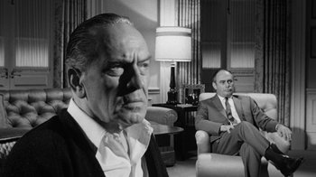 Movie still from “Seven Days in May” (1964), directed by John Frankenheimer – Two men sitting in a living room , one of whom is wearing a mask; Medium shot, Over the shoulder angle