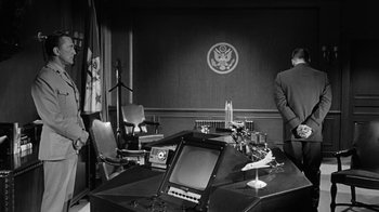 Movie still from “Seven Days in May” (1964), directed by John Frankenheimer – A man standing in front of a desk with a television; Wide shot, Low angle