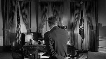 Movie still from “Seven Days in May” (1964), directed by John Frankenheimer – A black and white photo of a man standing in front of a desk; Medium shot, Over the shoulder angle