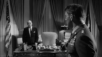 Movie still from “Seven Days in May” (1964), directed by John Frankenheimer – A man in a suit and tie standing in front of another man in a suit and tie in front of a desk; Medium shot, Over the shoulder angle