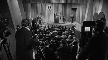 Movie still from “Seven Days in May” (1964), directed by John Frankenheimer – A crowd of people in a room watching a man speak; Wide shot, Over the shoulder angle