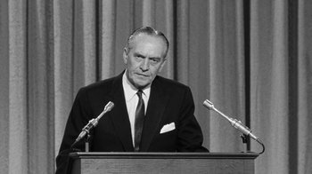 Movie still from “Seven Days in May” (1964), directed by John Frankenheimer – An older man in a suit and tie at a podium with two microphones; Medium shot, Low angle