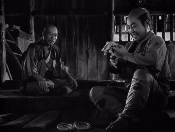 Movie still from “Seven Samurai” (1954), directed by Akira Kurosawa – Two men sitting on a bench in a room; Medium shot, Low angle