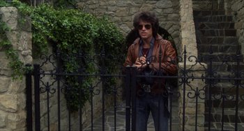 Movie still from “Shampoo” (1975), directed by Hal Ashby – A man standing next to a metal fence holding an umbrella; Medium shot, Low angle