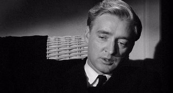 Movie still from “Ship of Fools” (1965), directed by Stanley Kramer – A man in a suit and tie sitting in front of a wicker chair; Close Up shot, Over the shoulder angle