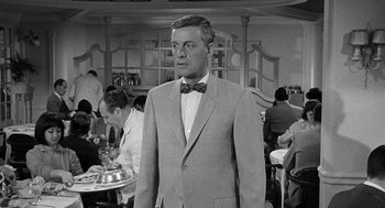 Movie still from “Ship of Fools” (1965), directed by Stanley Kramer – A man in a suit and bow tie standing in front of a table; Medium shot, Over the shoulder angle