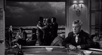 Movie still from “Ship of Fools” (1965), directed by Stanley Kramer – A man sitting at a table in front of a group of people; Wide shot, Over the shoulder angle