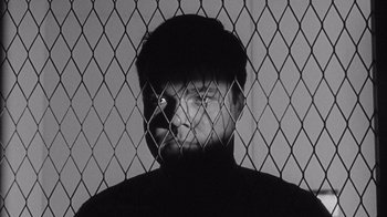 Movie still from “Shock Corridor” (1963), directed by Samuel Fuller – A man is behind a chain link fence looking at the camera; Close Up shot, Low angle