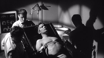 Movie still from “Shock Corridor” (1963), directed by Samuel Fuller – A man is strapped to a chair in a room with two other men; Medium shot, High angle