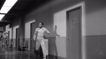 Movie still from “Shock Corridor” (1963), directed by Samuel Fuller – Black and white photograph of a man walking in a hallway; Wide shot, High angle