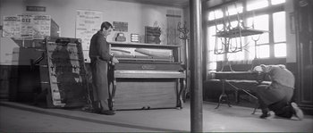 Movie still from “Shoot the Piano Player” (1960), directed by François Truffaut – A man standing in front of an upright piano in a room; Wide shot, High angle